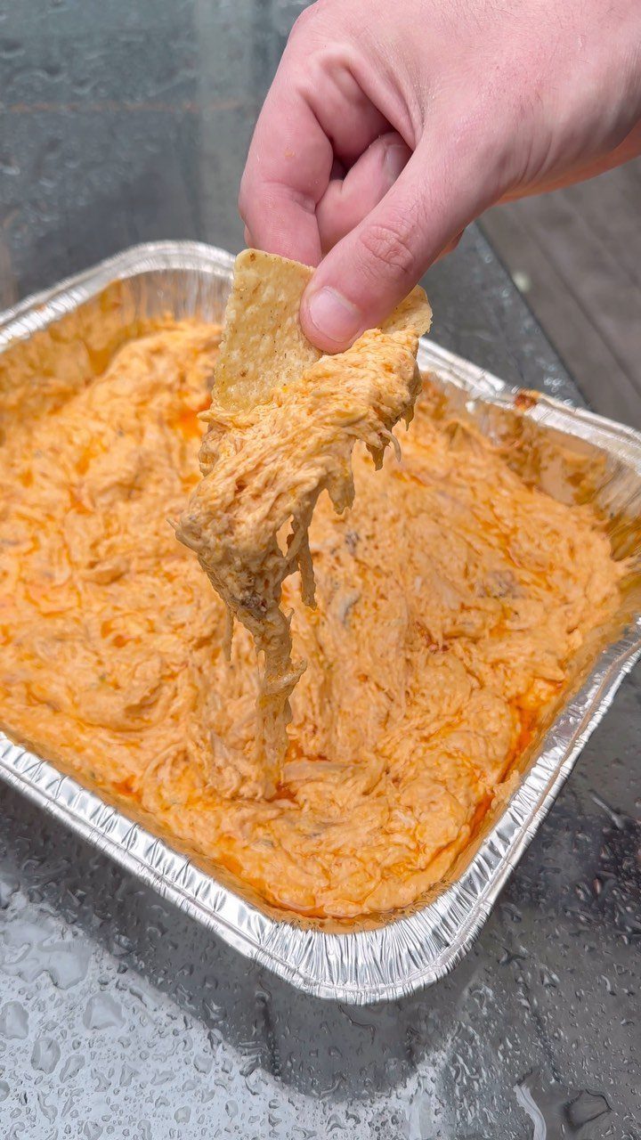 Tortilla chip scooping creamy orange buffalo chicken dip from an aluminum foil tray.