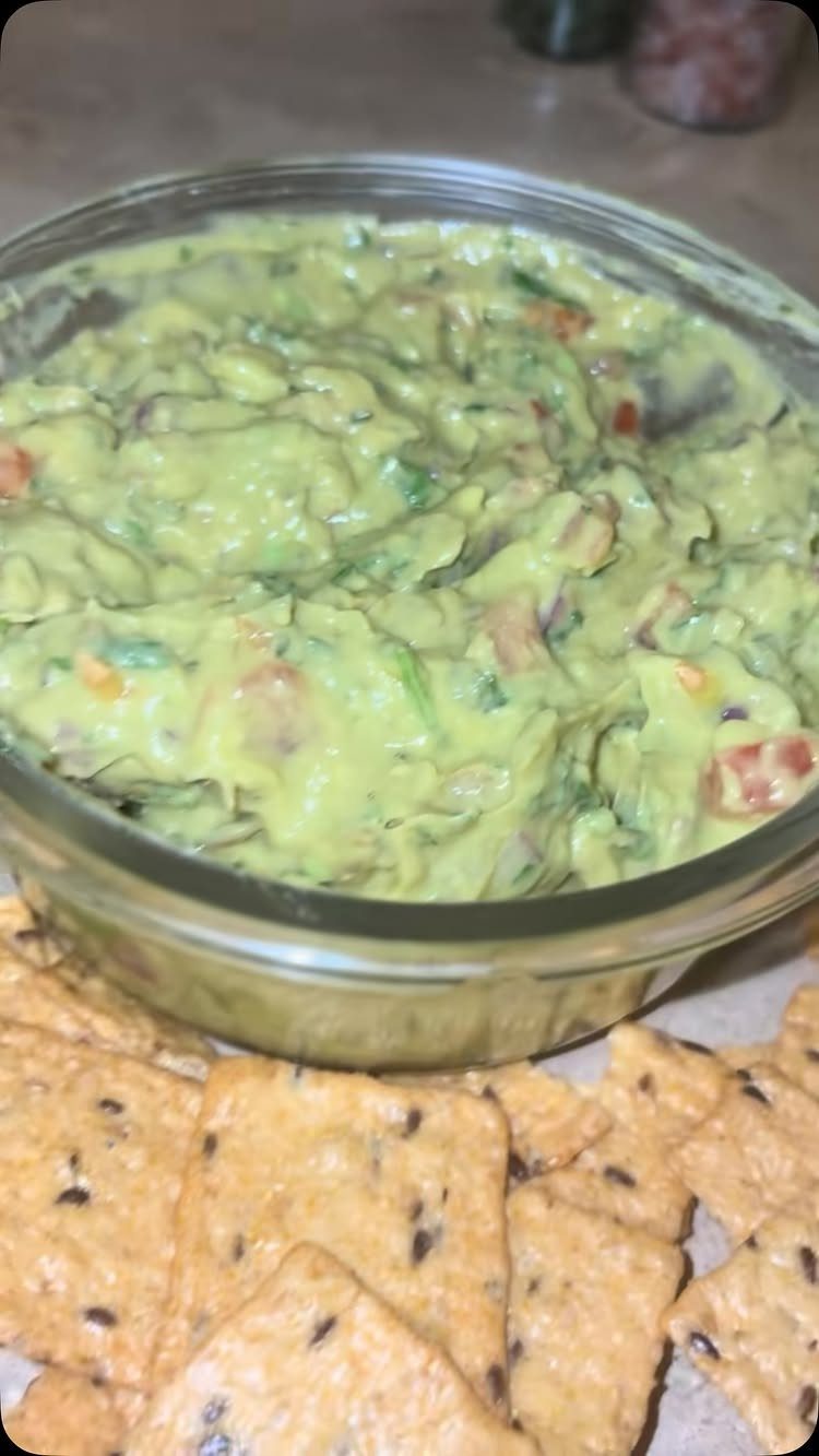 Guacamole in a glass bowl served with square seeded crackers on a table.