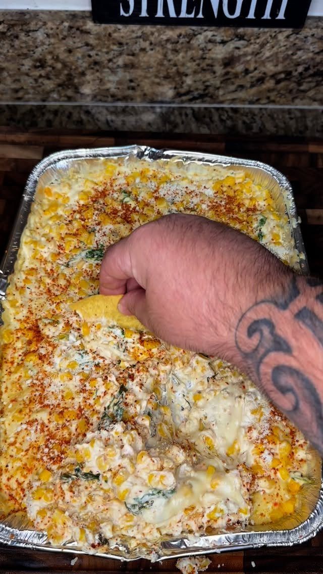 Hand dipping a tortilla chip into a cheesy corn dip in a foil pan.