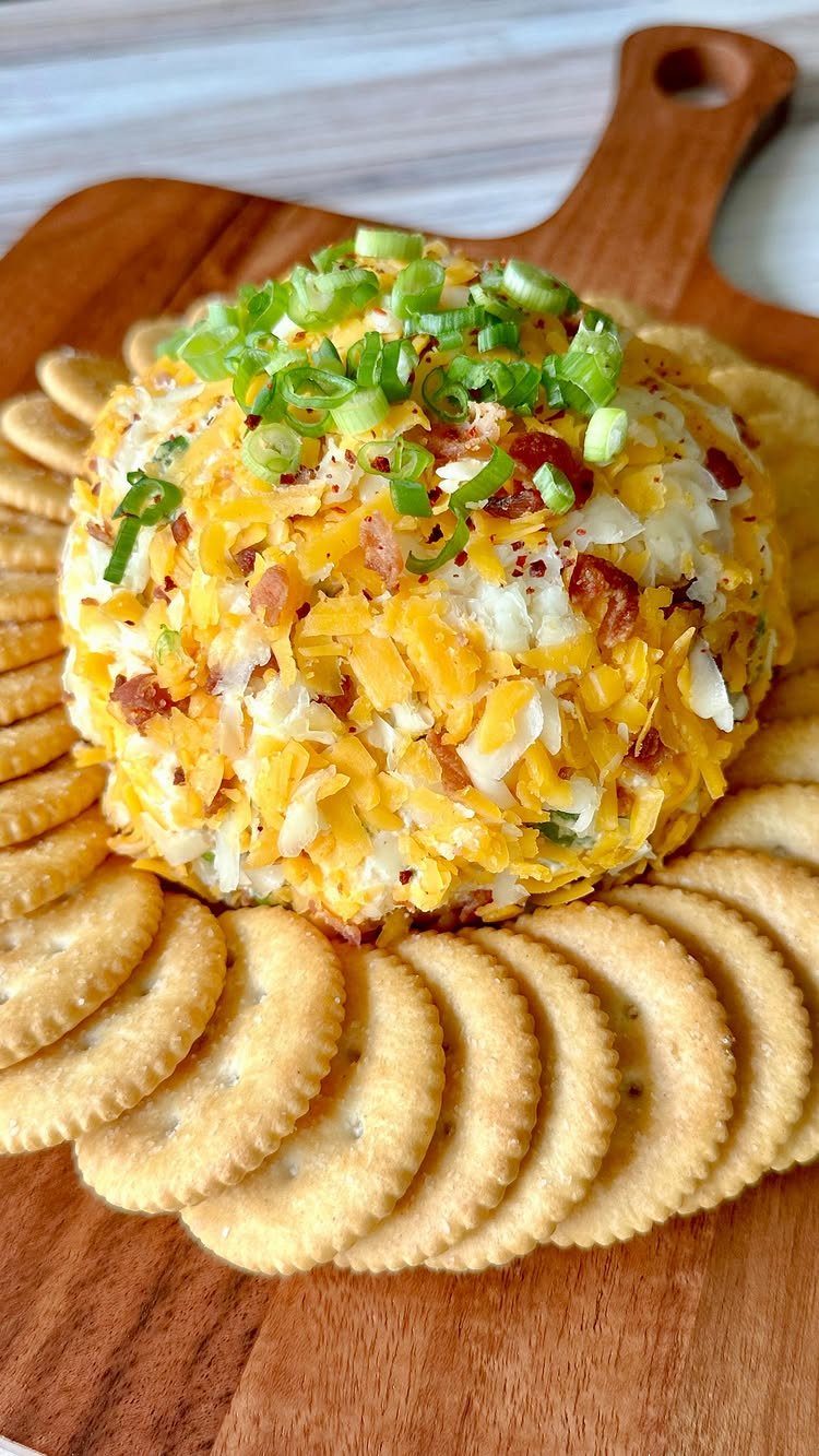 Cheese ball topped with green onions and bacon, surrounded by crackers on wood.