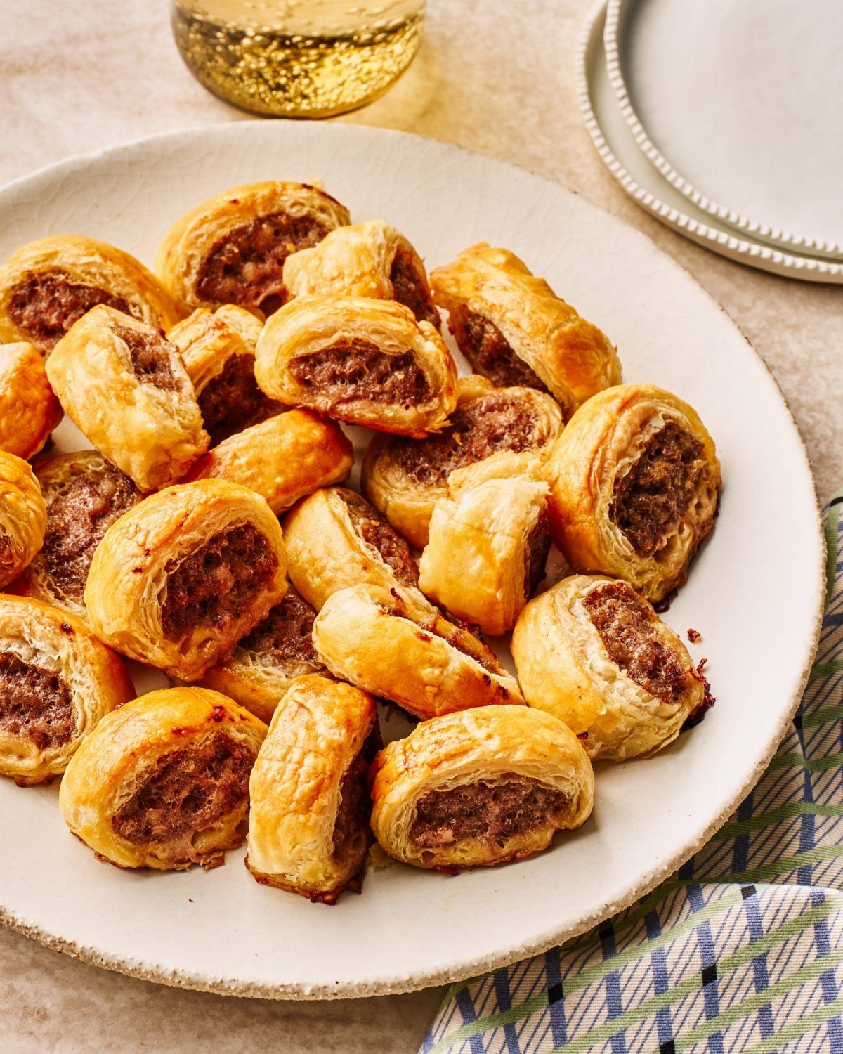 Golden-brown puff pastry sausage rolls piled on a white ceramic plate.