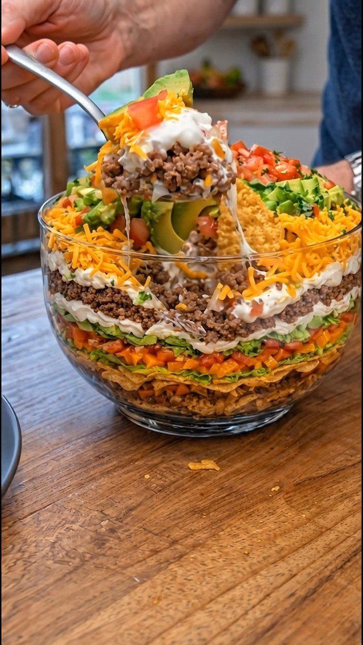 Seven-layer dip in a glass bowl with a scoop being lifted out.