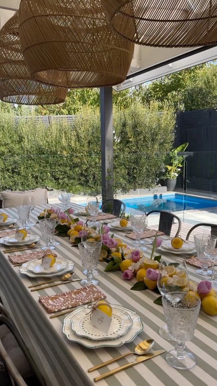 Outdoor dining table decorated with lemons, pink flowers, and striped tablecloth near a pool.