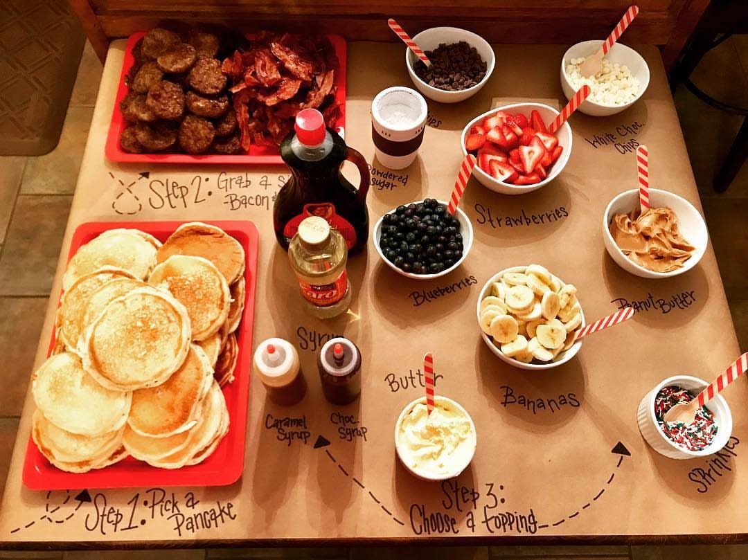 Pancake bar with toppings, syrups, and breakfast meats on a brown paper table.