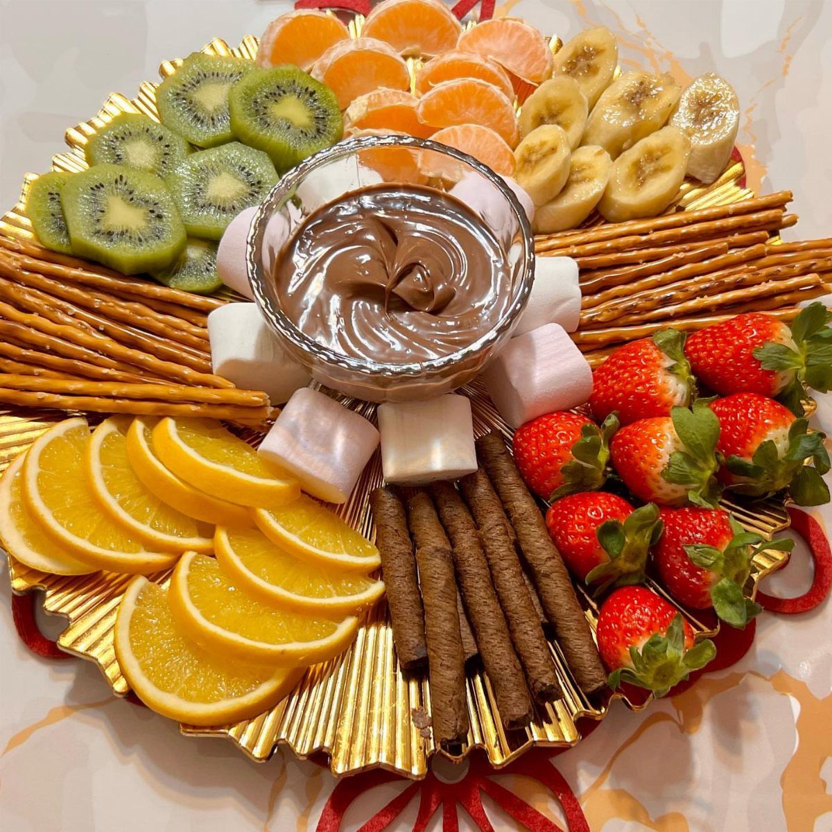 Chocolate dip bowl surrounded by fresh fruit, marshmallows, and pretzels on gold platter.
