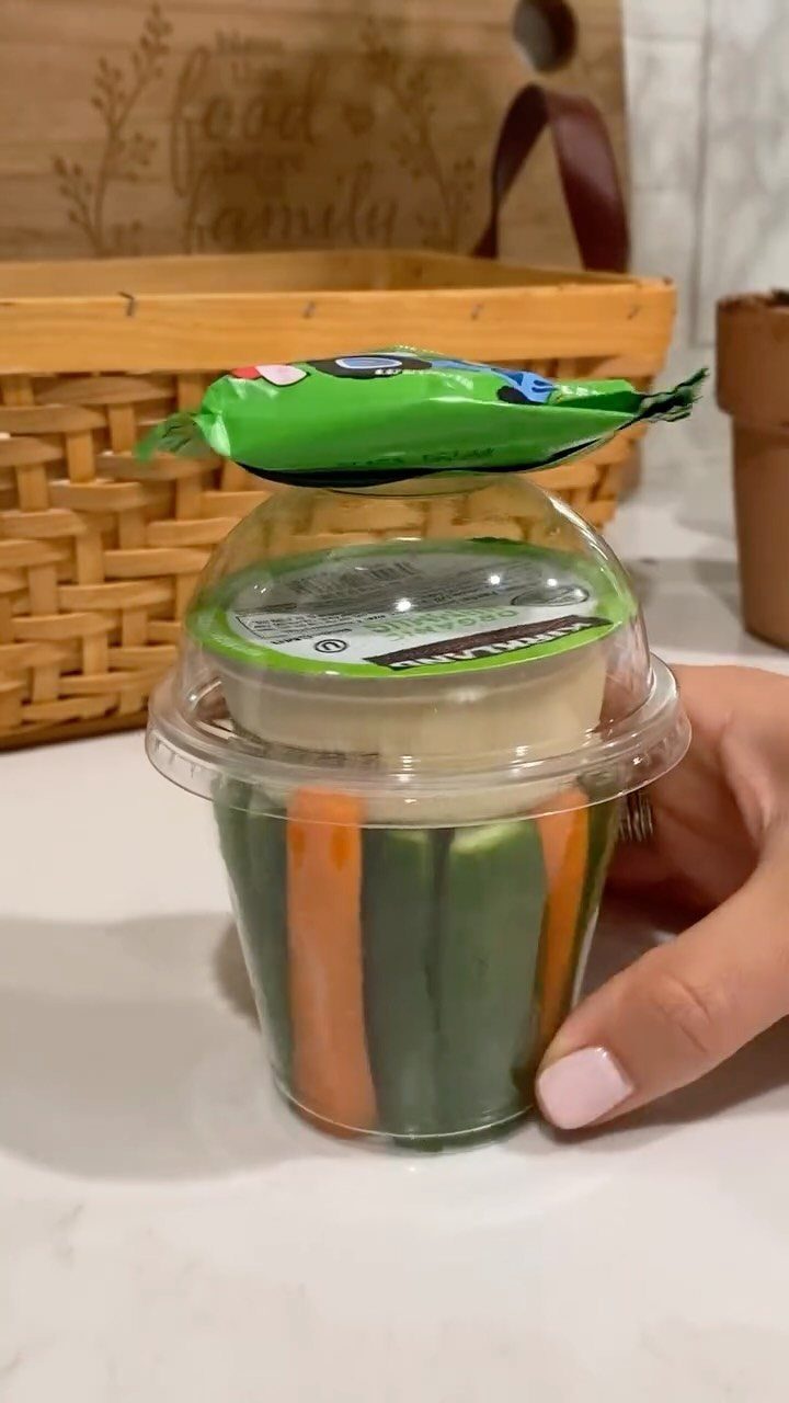 Hummus dip cup with carrot and cucumber sticks held by a hand.