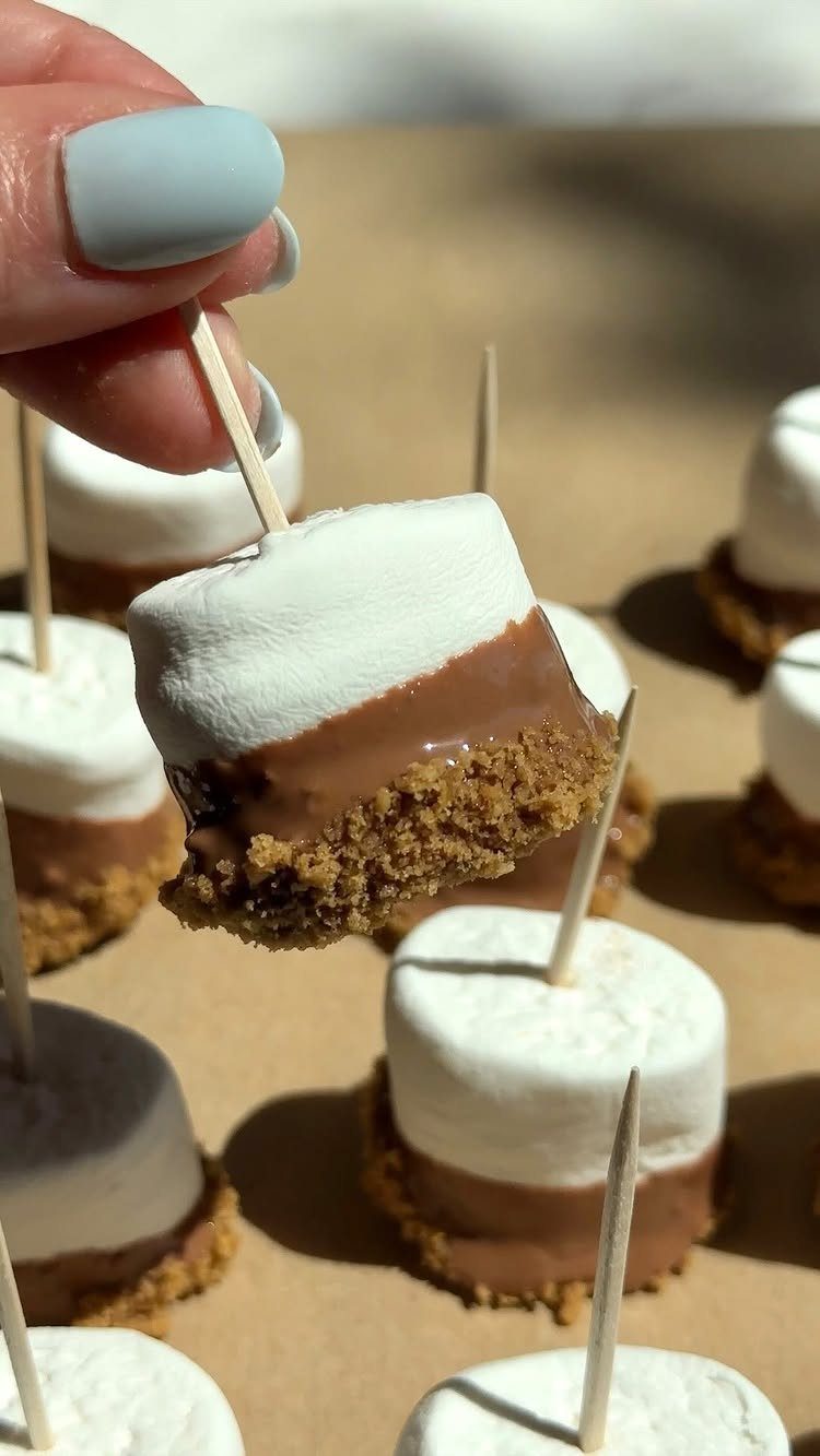 S'more bites with graham cracker, chocolate, and marshmallow on toothpicks held by hand.