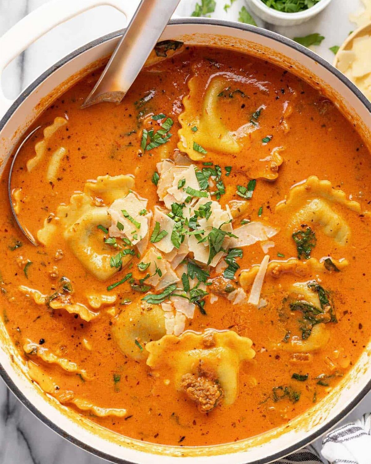 Creamy tomato ravioli soup in a white pot topped with parmesan and parsley.