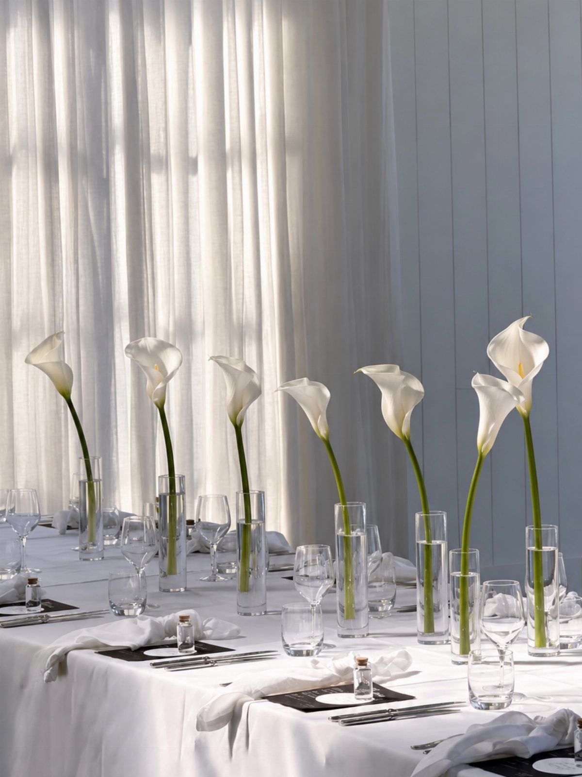White calla lilies in slender glass bud vases lining a white dining table.