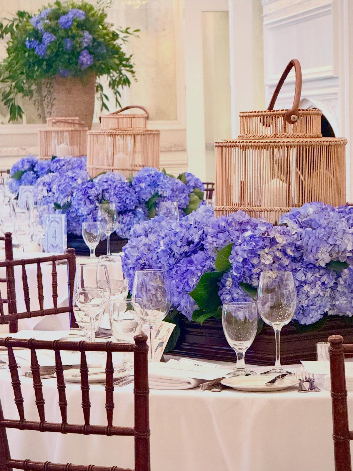 Blue hydrangea floral arrangements and woven lanterns on a white dinner table.