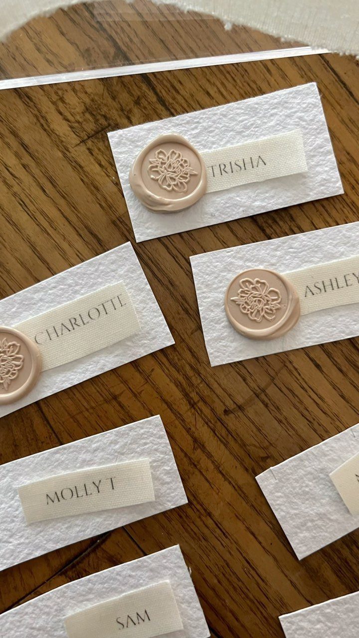 White textured place cards with gold names and beige floral wax seals on wood.