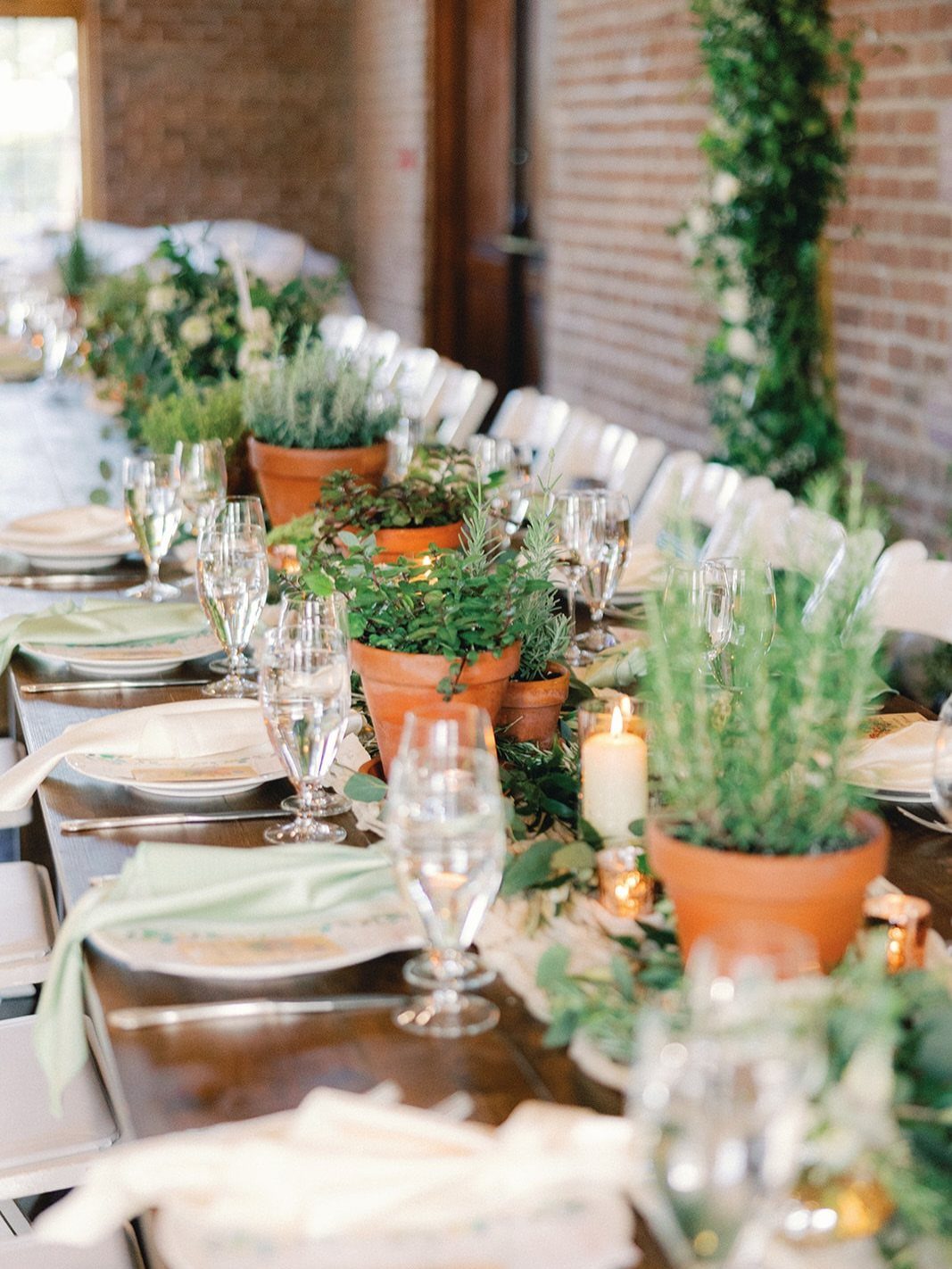 Terracotta pots with green herbs and white candles as centerpieces on a wooden table.