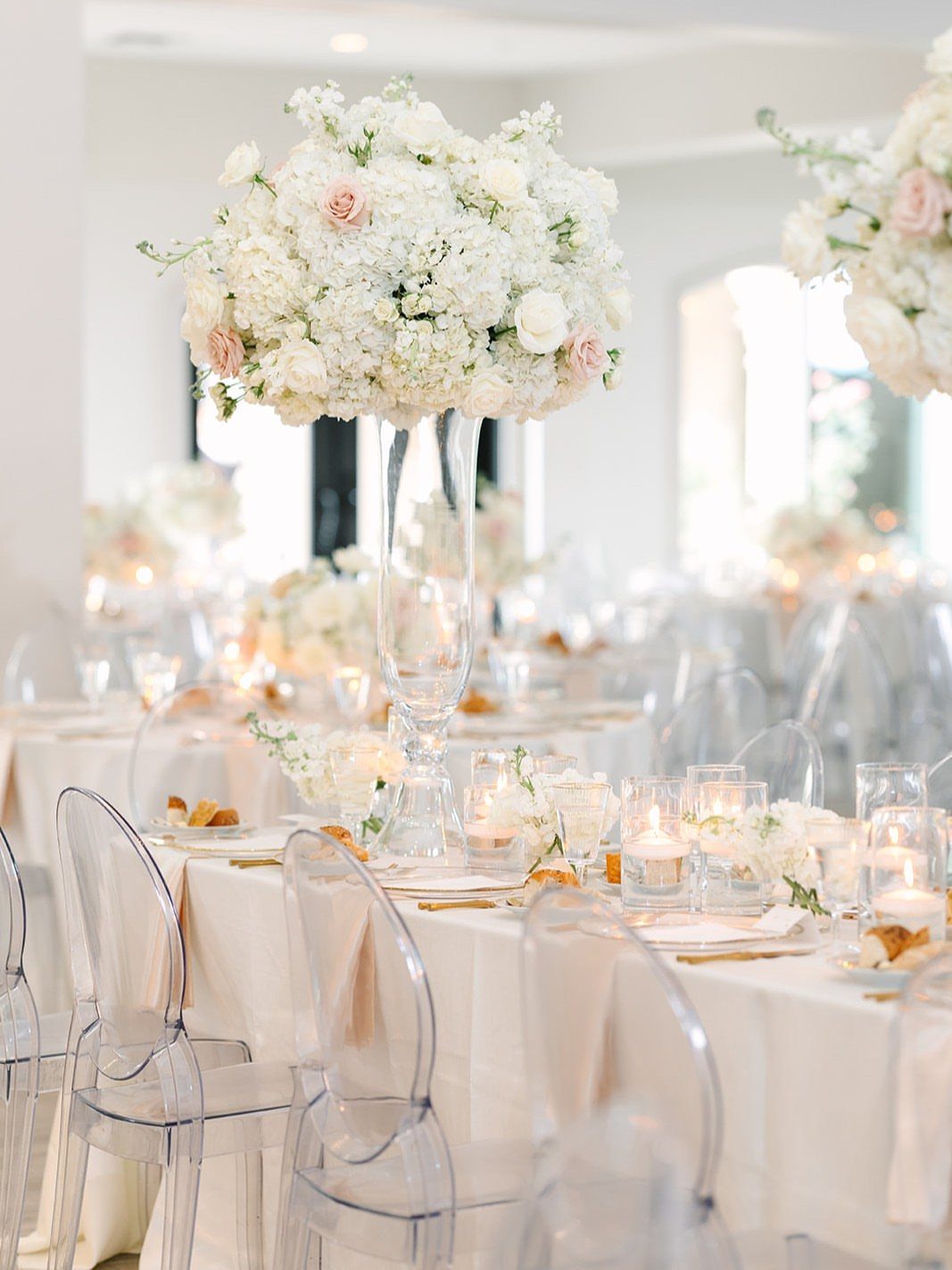 Tall white floral centerpiece and small candles on a white dinner table.