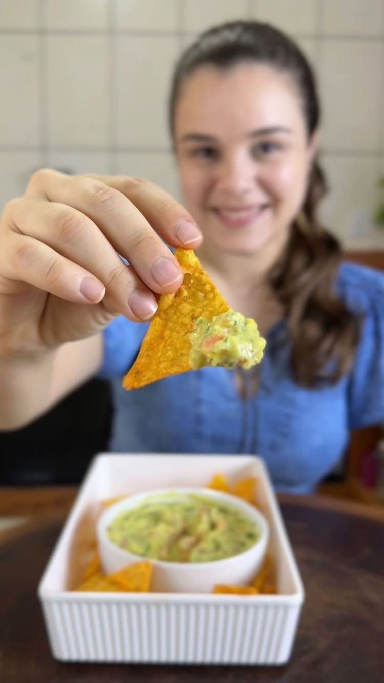 Tortilla chip dipped in green guacamole held by a smiling woman in kitchen