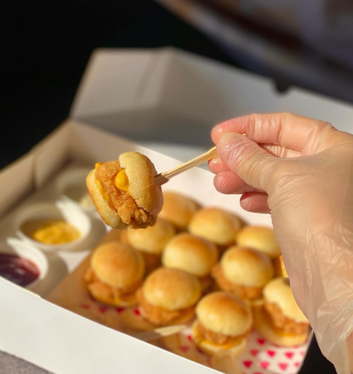 Mini chicken burgers with cheese in a white box, one held by a hand.