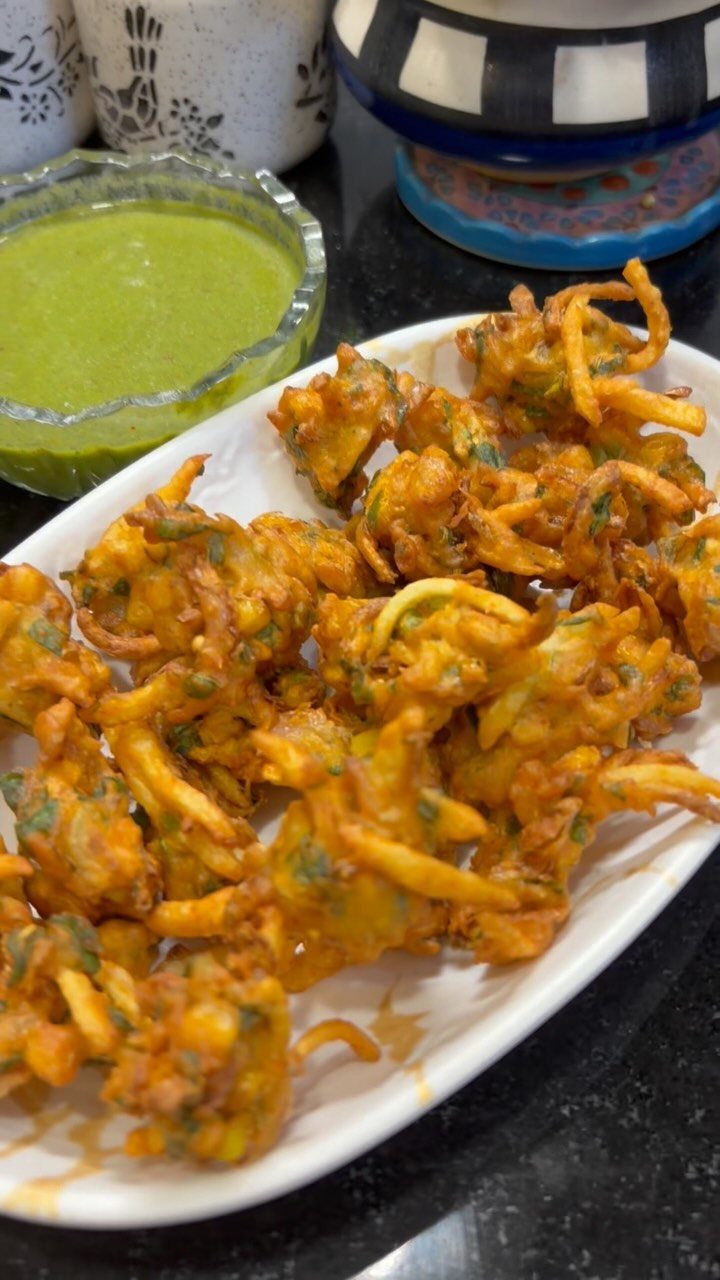 Golden-brown vegetable pakoras on a white platter with a side of green chutney.