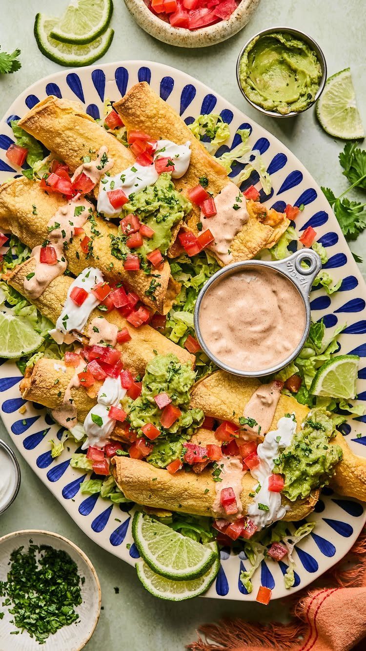Baked chicken taquitos topped with guacamole, tomatoes, and cream on a patterned platter.