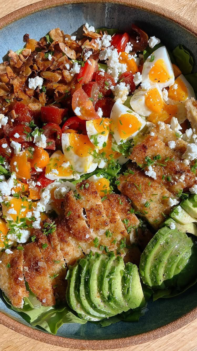 Grilled chicken salad with avocado, boiled eggs, tomatoes, and crumbled cheese in a bowl.