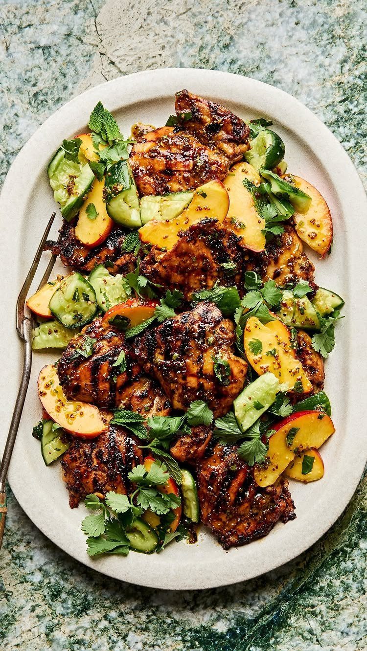 Grilled chicken with sliced peaches, cucumbers, and fresh herbs on a white platter.