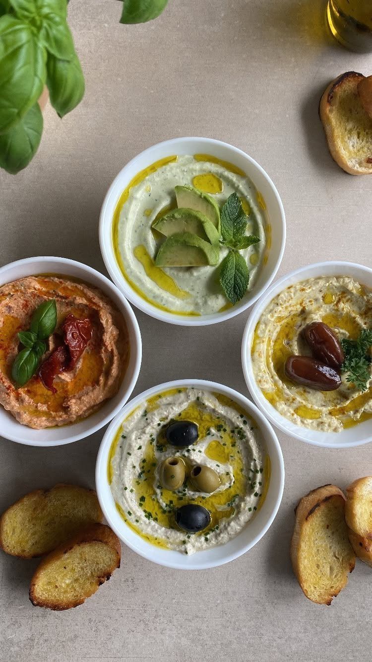 Four bowls of colorful dips with toasted bread and fresh basil leaves.