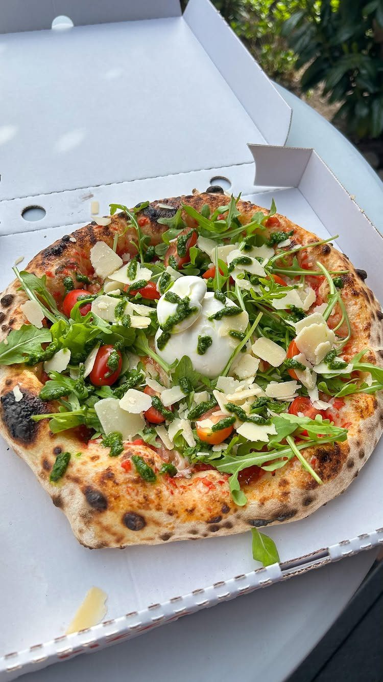 Pizza with arugula, cherry tomatoes, and shaved parmesan in a white delivery box.
