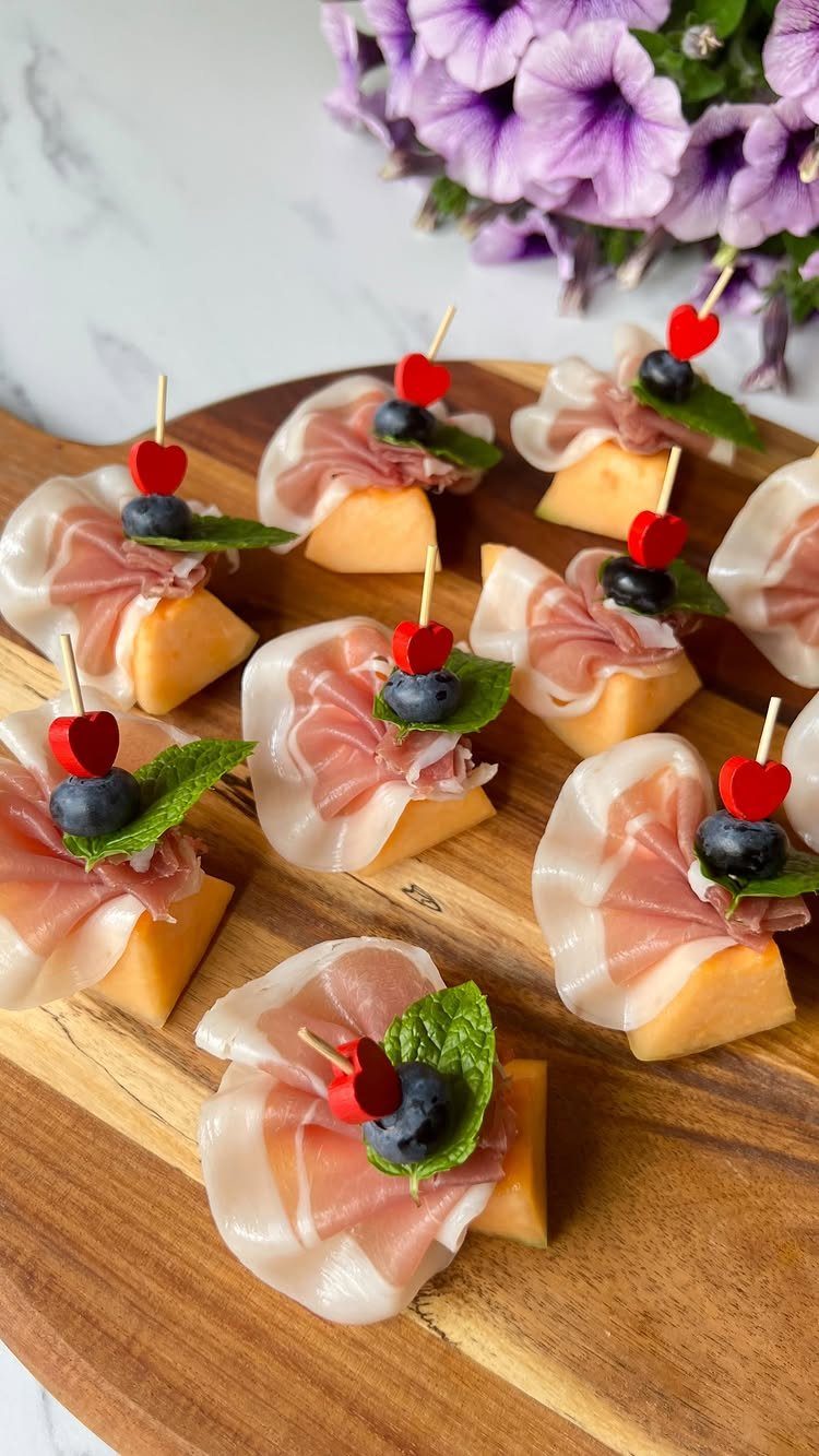 Prosciutto and cantaloupe appetizers with blueberries and mint on a wooden board.