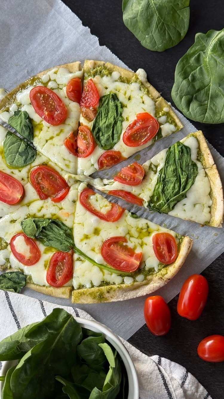 Pesto pizza with melted mozzarella, cherry tomatoes, and fresh spinach on parchment paper.