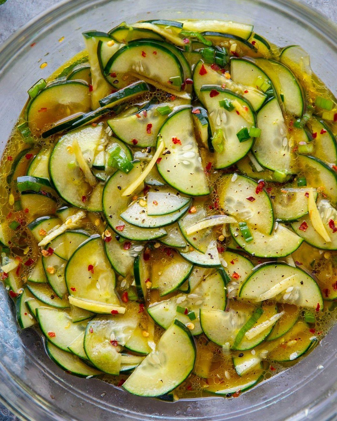 Sliced cucumbers with ginger, red pepper flakes, and green onions in a glass bowl.