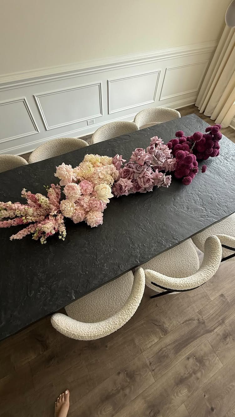 Elongated floral arrangement of pink and purple flowers on a dark rectangular table.
