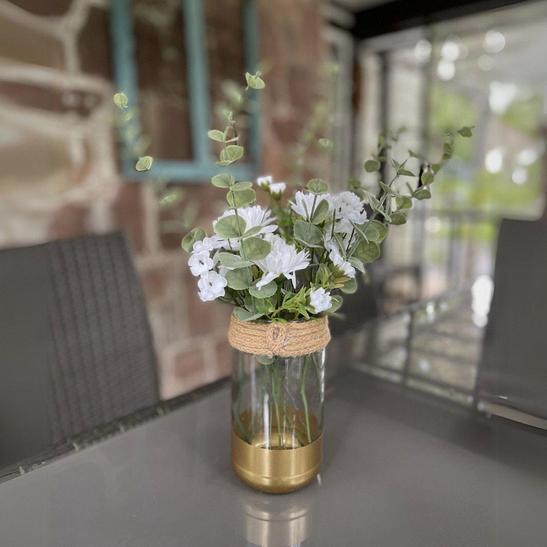 Glass jar vase with gold base and twine holding white flowers and greenery.