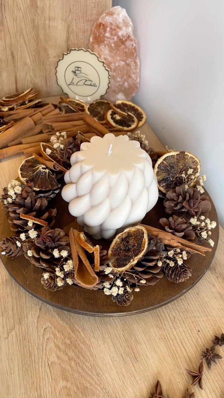 White textured pillar candle surrounded by pinecones and dried citrus on wooden platter