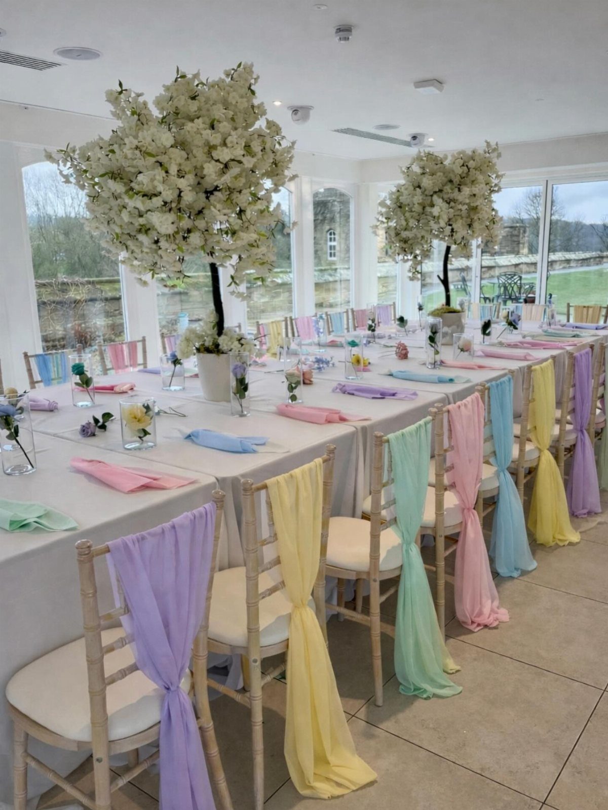 Long white dinner table with pastel fabric chair sashes and white floral centerpieces.