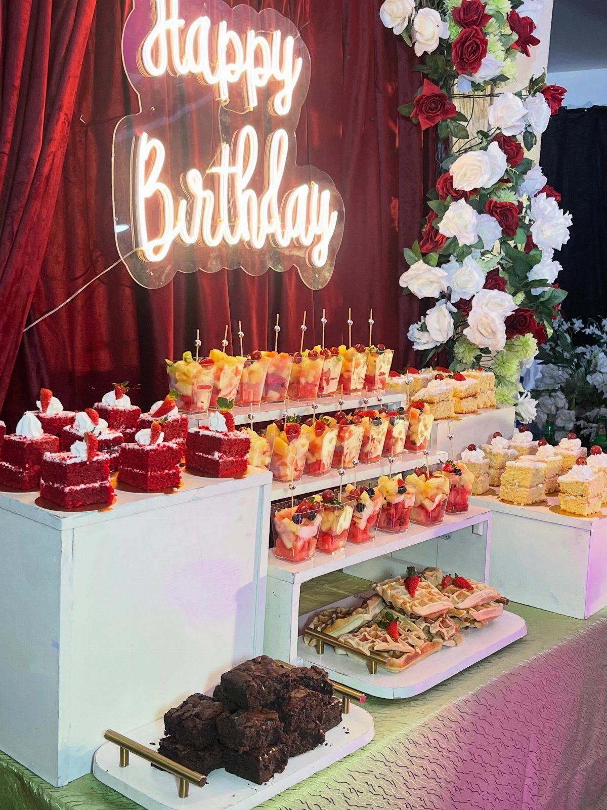 Dessert buffet table with cakes and parfaits under a neon Happy Birthday sign.