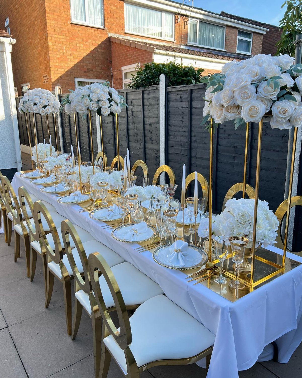Long white table with gold chairs and tall white rose centerpieces outdoors.