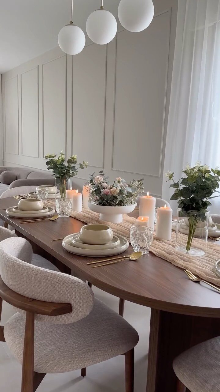 Wooden dining table with beige dinnerware, gold cutlery, white candles, and floral centerpieces.