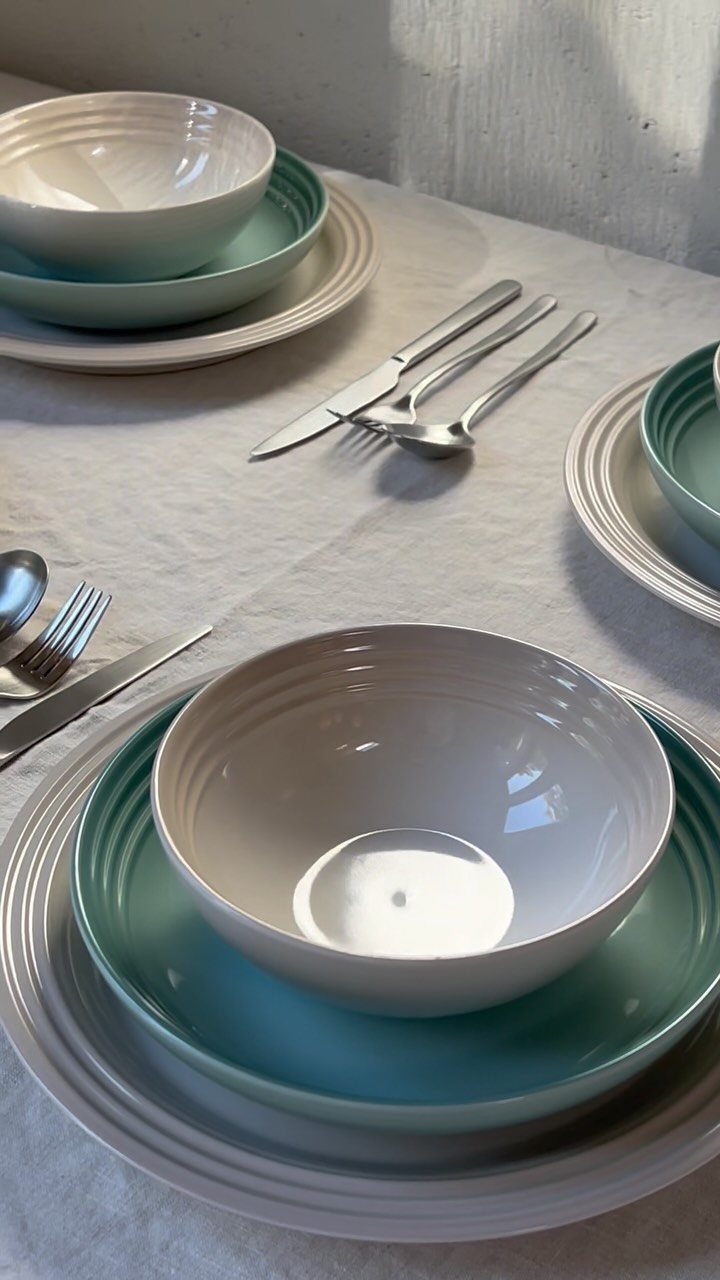 Stacked white and teal plates and bowls with silver cutlery on white tablecloth.