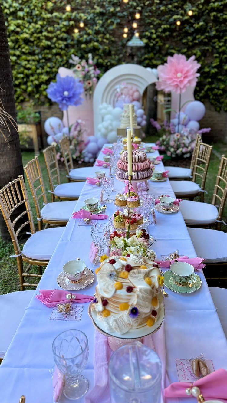 Pastel tea party table with pink napkins, macarons, and a decorated cake outdoors.