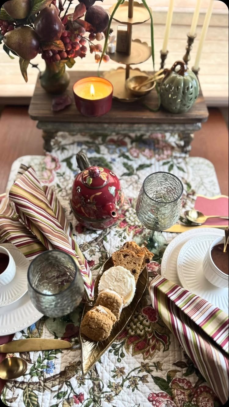 Tea party table with autumn fruits, red teapot, and pastries on floral linens.