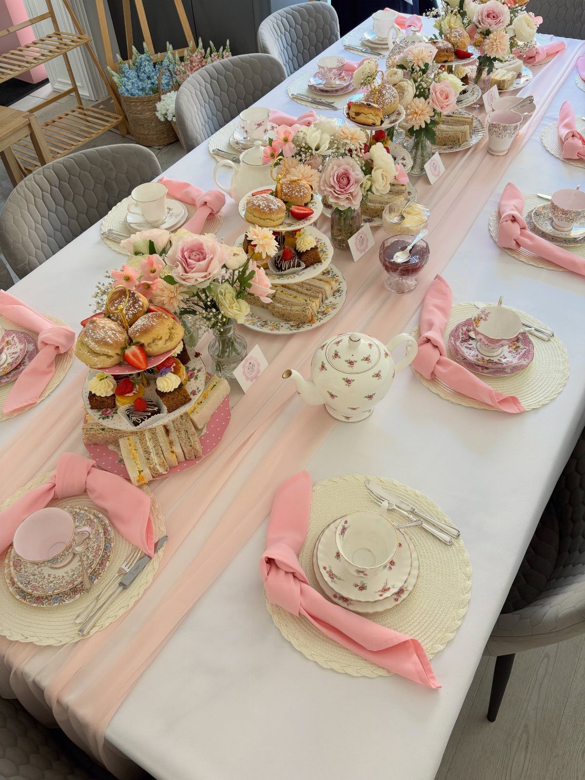 Tea party table set with pink linens, floral porcelain, tiered treats, and roses.