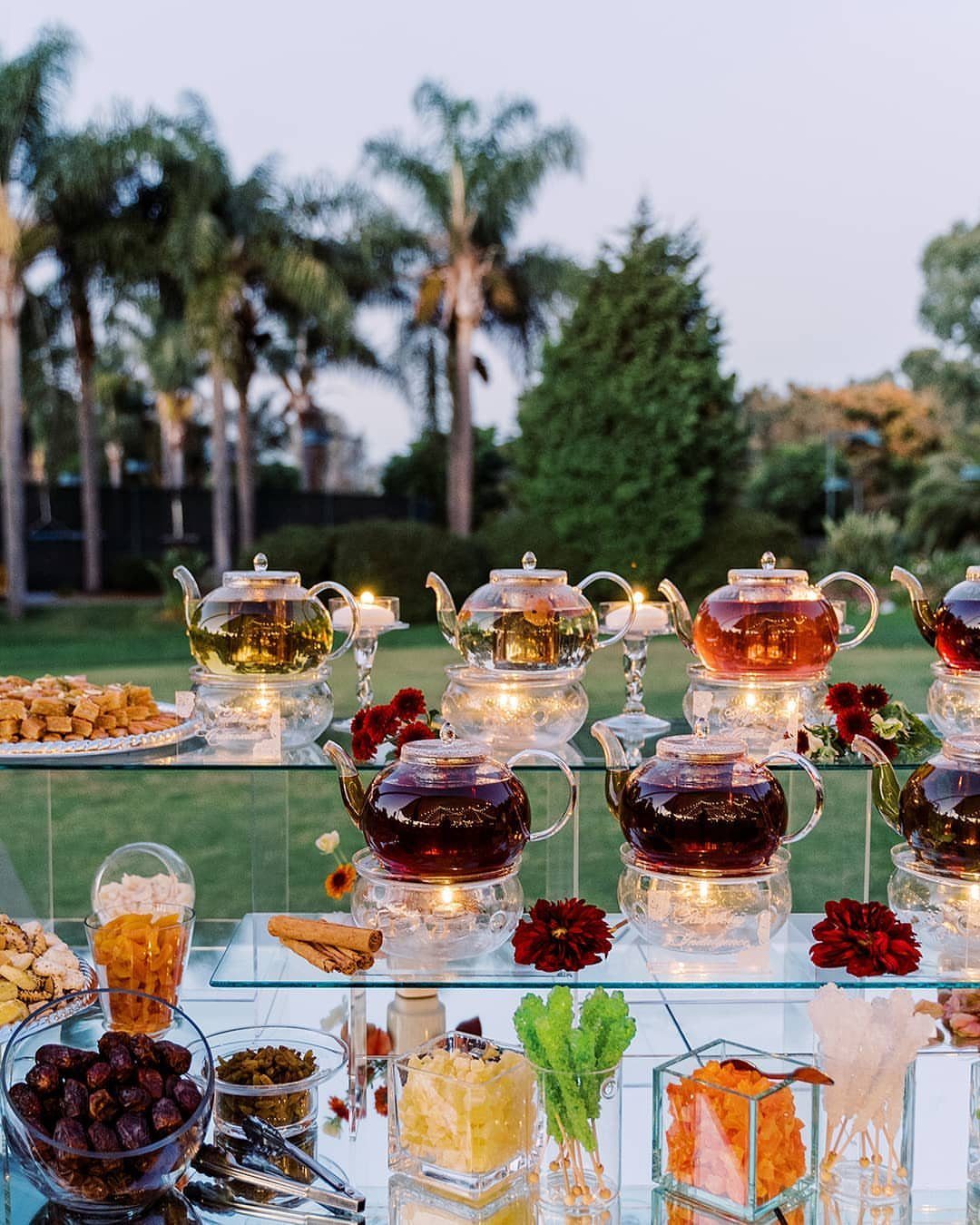 Glass teapots and lit candles on a table with snacks in a garden.