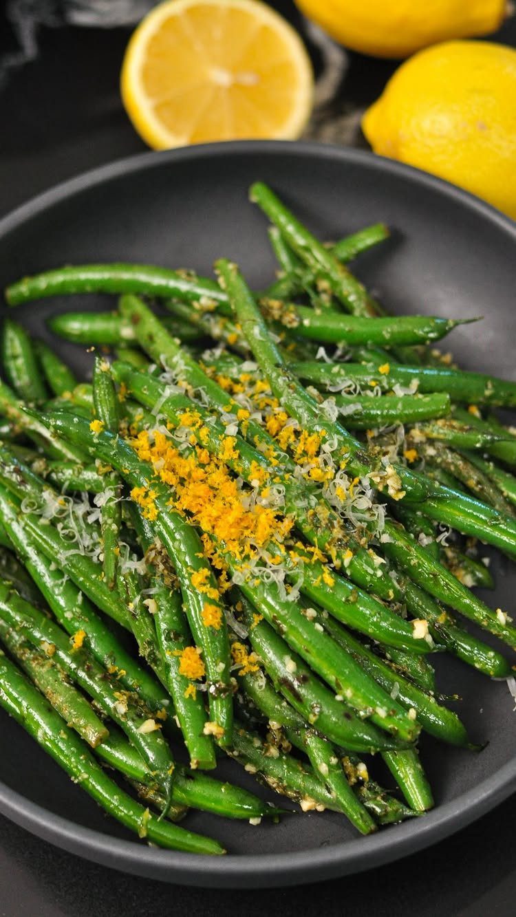 Green beans with lemon zest and cheese in a black bowl with lemons.