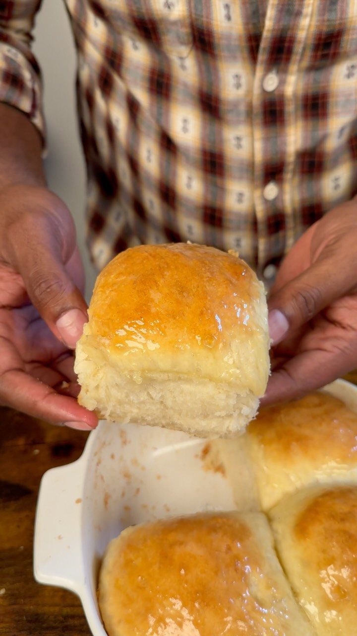Golden-brown glazed dinner roll held above a white baking dish of rolls.
