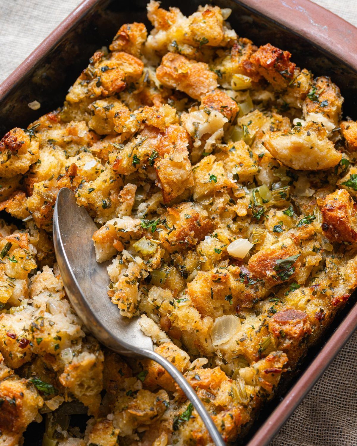 Golden-brown bread stuffing with herbs in a baking dish with a metal spoon.