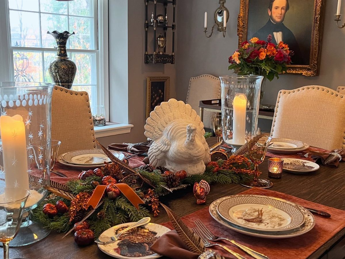 Thanksgiving dining table with white turkey centerpiece, evergreen greenery, and autumn-themed plates.