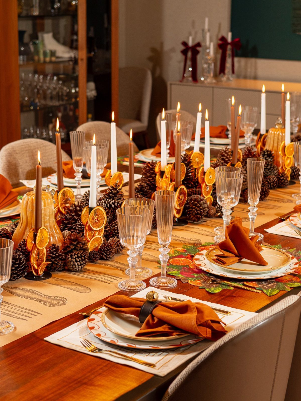 Thanksgiving table setting with orange napkins, taper candles, pinecones, and dried orange slices.