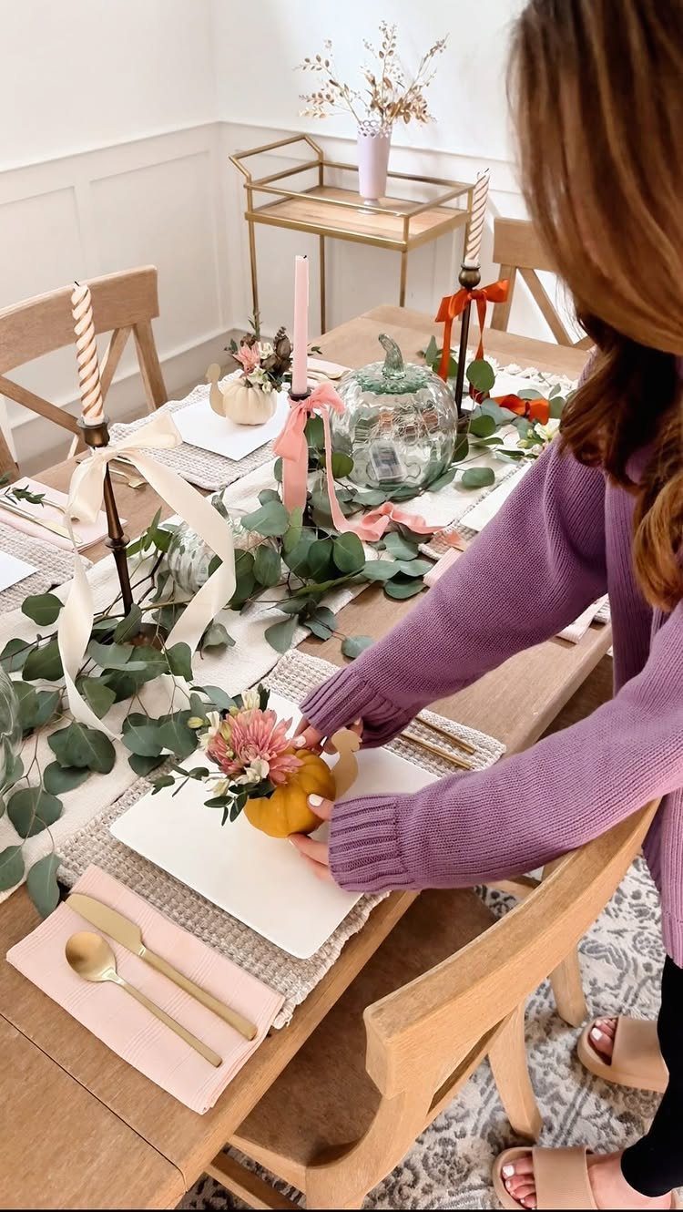 Woman arranging a Thanksgiving table with gold cutlery, greenery, and decorative pumpkins.
