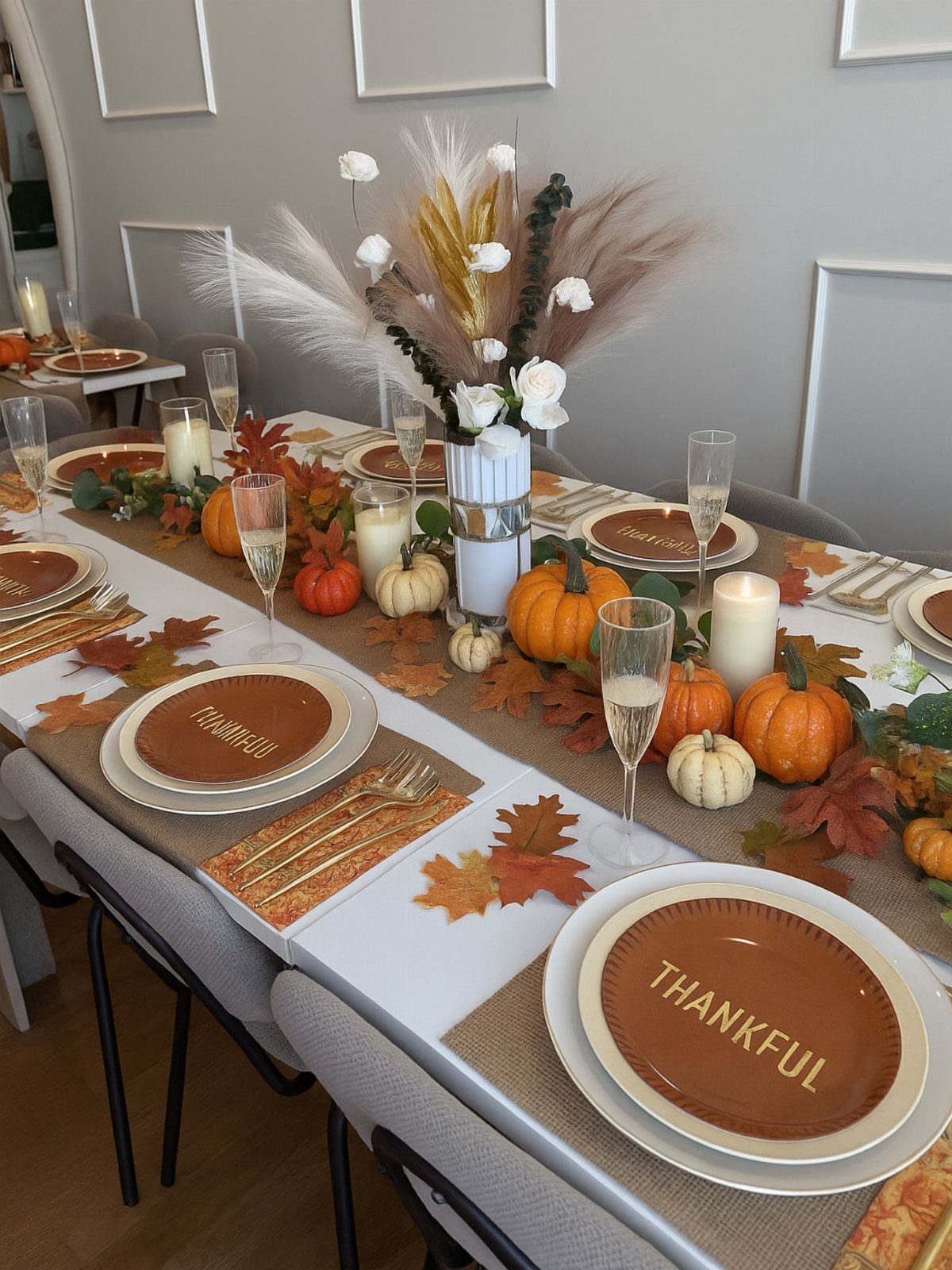 Thanksgiving table setting with pumpkins, pampas grass, gold cutlery, and white pillar candles.