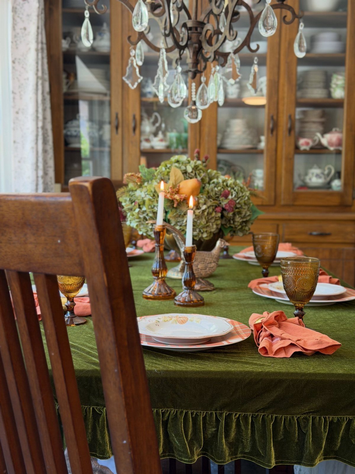 Green velvet tablecloth with orange napkins, amber glasses, and lit candles for Thanksgiving.