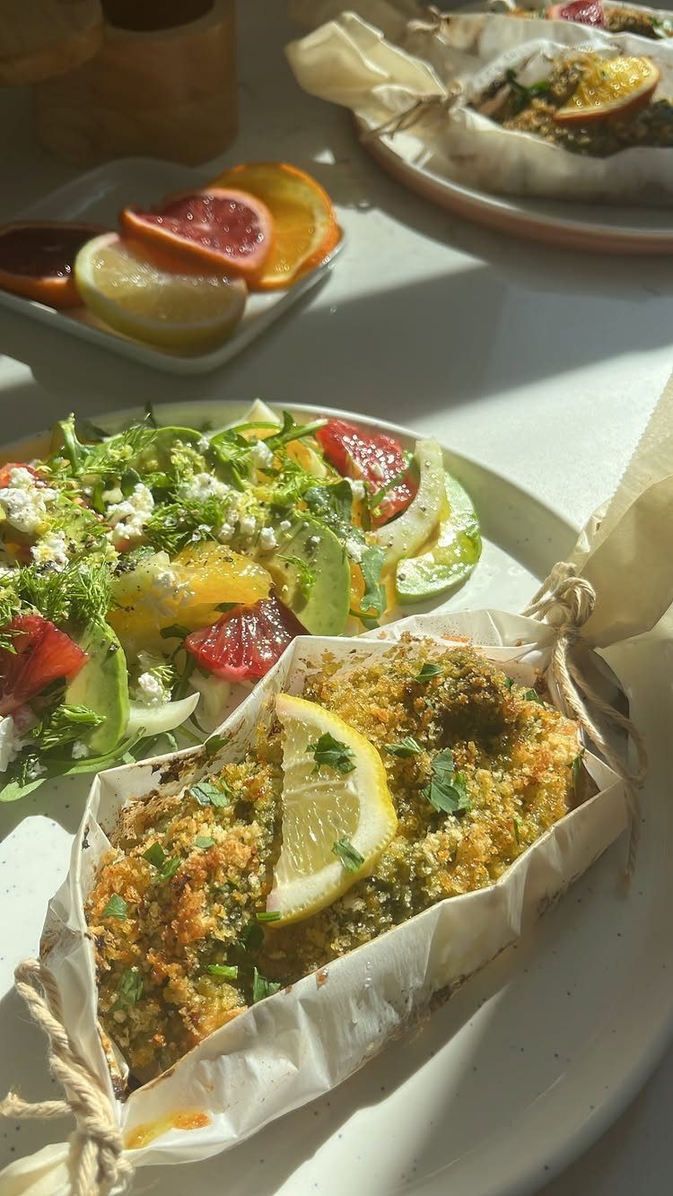 Breaded fish fillet with lemon in parchment paper next to a citrus salad.