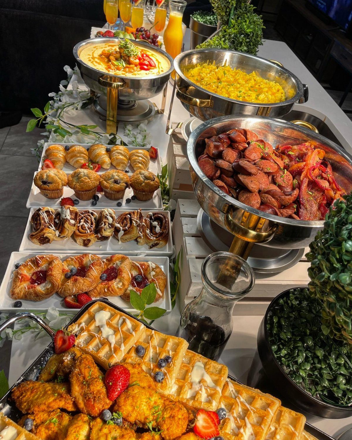 Breakfast buffet spread with waffles, pastries, scrambled eggs, and sausages on a white table.